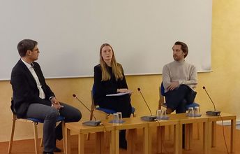 Drei Personen sitzten an Sprecherpulten uns unterhalten sich miteinander.