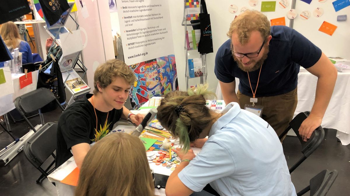 Zwei junge Menschen malen etwas auf ein Papier, ein Mann steht daneben und schaut zu