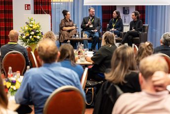 Vier Personen sitzen auf einem Podium und diskutieren während das Publikum zuhört.