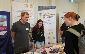 Zwei Personen stehen an einem Infostand und unterhalten sich mit einer Frau