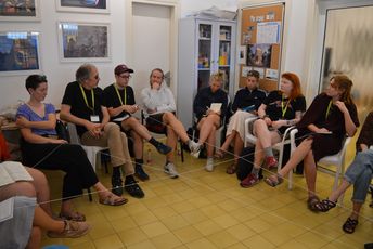 Eine Gruppe von Menschen sitzt in einem Sruhlkreis, der von Fäden durchkreuzt wird, die die Teilnehmenden in der Hand halten.