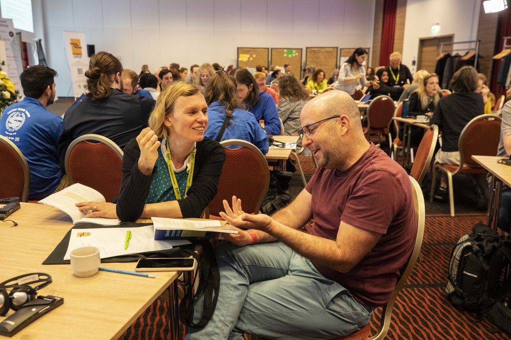 Zwei Menschen im angeregten Gespräch zu vor ihnen liegendem Buch während einer Tagung.