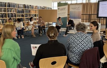 Es ist eine Seminarsituation zu sehen, in der in einem Stuhlkreis die Teilnehmenden in Kleingruppen etwas besprechen. Die Referent*innen sitzen im Hintergrund vor zwei Werbebannern der Veranstalter.