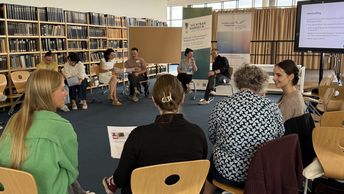 Es ist eine Seminarsituation zu sehen, in der in einem Stuhlkreis die Teilnehmenden in Kleingruppen etwas besprechen. Die Referent*innen sitzen im Hintergrund vor zwei Werbebannern der Veranstalter.