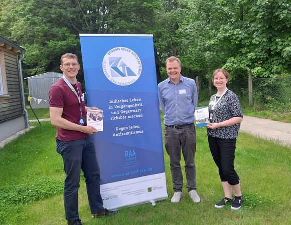 Drei Personen stehen vor einem Banner vom Bündnis gegen Antisemitismus