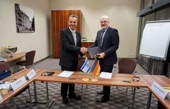 Zwei Männer schütteln Hände. Auf dem Tisch vor ihnen stehen die Deutsche unf Israelische Landesflagge.