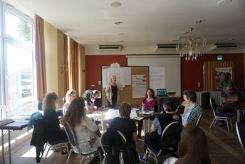 Die Teilnehmenden sitzen in einem Raum und hören einer Frau zu, die vorne steht und zu der Gruppe spricht.