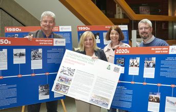 Vier Menschen lächeln in die Kamera und halten blaue und weiße Ausstellungstafeln der Präsentation zu 70 Jahre deutsch-israelischem Jugendaustausch hoch.
