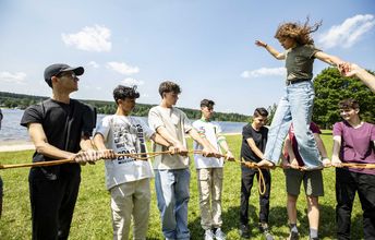 Eine junge Frau balanciert auf einem Seil, welches von anderen Jugendlichen gespannt wird. Im Hintergrund sind ein See und Wald.