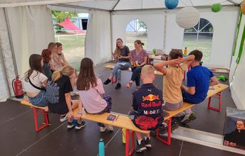 Jugendliche sitzen auf Bänken in einem Zelt und hören zwei Teamerinnen gespannt zu.