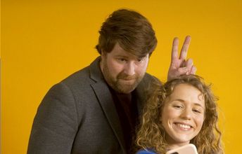 Eine Frau macht ein Selfie, im Hintergrund steht ein Mann und macht Hasenohren - Werbebild für die Online-Plattform DINA.international