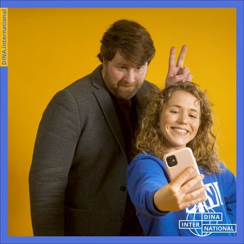 Eine Frau macht ein Selfie, im Hintergrund steht ein Mann und macht Hasenohren - Werbebild für die Online-Plattform DINA.international