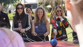 Drei Frauen stehen auf einer Wiesen und ziehen an Bändern, auf einem Tisch liegt eine Kugel, die hin und her balanciert wird