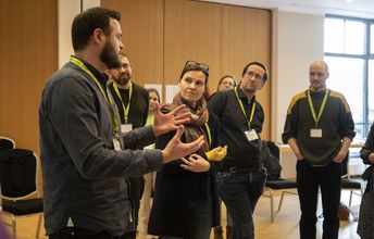 Eine stehende Gruppe von Seminarteilnehmenden hört konzentriert einem der Teilnehmenden zu, der gerade spricht udn etwas erläutert.