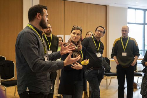 Eine stehende Gruppe von Seminarteilnehmenden hört konzentriert einem der Teilnehmenden zu, der gerade spricht udn etwas erläutert.