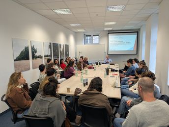 Gruppe sitzt um einen Tisch und hört einem Referenten interssiert zu, hinter dem eine Präsentation aufgerufen ist.