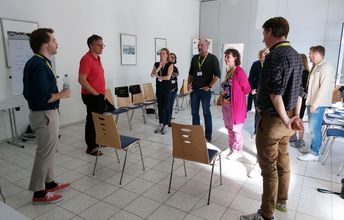 Stehende Seminarteilnehmende in einem interessierten Gespräch mit dem Moderator, zwischen Stühlen stehend.