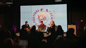 Drei Personen sitzen vor einem erleuchteten Hintergrund in einem dunklen Raum auf einem Podium