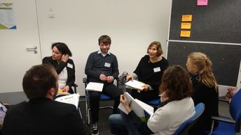 Eine Kleringruppe sitzt diskutierend im Kreis; an einer Pinnwand hängen gesammelte Notizzettel.