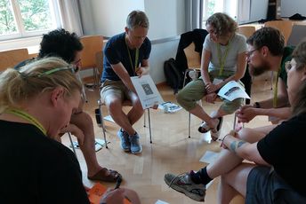 Kleingruppe sitzt in einem Stuhlkreis und bespricht konzentriert Arbeitsmaterialien mit Text und Bild.