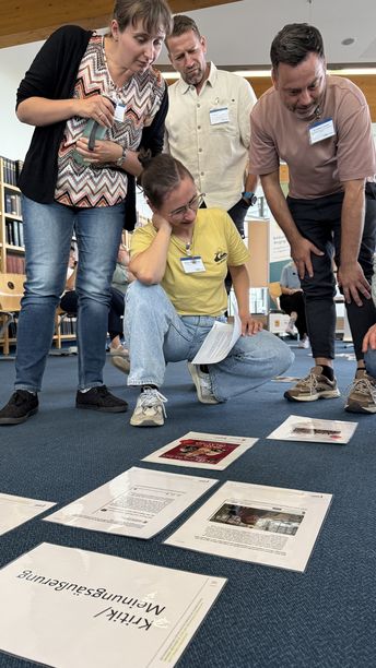 Eine Gruppe von vier Menschen betrachtet hockend und sitzend Bild- und Textmaterial, das auf dem Boden ausgebreitet ist.