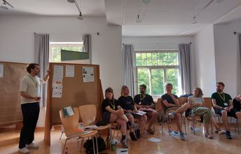 Eine im Halbrund sitzende Gruppe hört einem Moderator zu, der etwas an einer Pinnwand stehend erklärt. (Seminarsituation)