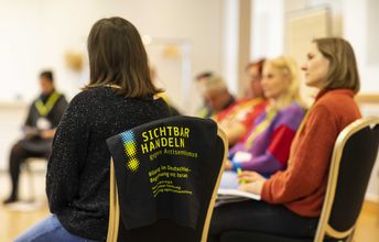 Seminargruppe sitzt im Stuhlkreis, im Vordergrund ist ein schwarzer Jutebeutel mit dem Aufdruck des Projekttitels "Sichtbar handeln! Gegen Antisemitismus." zu sehen.