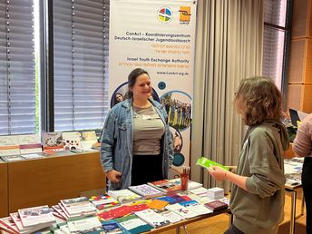 Eine Frau steht an einem Infostand mit Materialien und spricht mit einer Person