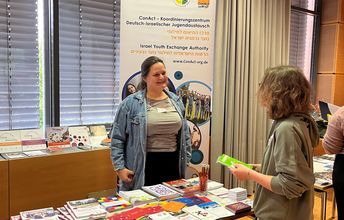 Eine Frau steht an einem Infostand mit Materialien und spricht mit einer Person