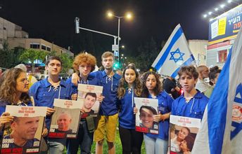 Gruppe Jugendlicher in blauen Hemden (israelische Jugendorganisation HaShomer Hatzair) demonstrieren und halten Plakate mit Fotos, Namen und Alter der Entführten vom 7.10.2023 hoch. Im Hintergrund sind israelische Fahnen zu sehen.